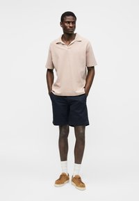 Ein Mann steht mit den Händen in den Taschen, trägt ein beiges kurzärmliges Poloshirt, marineblaue Shorts, weiße Socken und braune Freizeitschuhe.