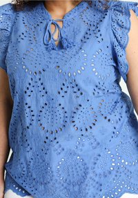 Blusa azzurro chiaro con pattern floreali ricamati e disegni a intaglio. Presenta maniche corte arricciate e una scollatura con lacci e frange.