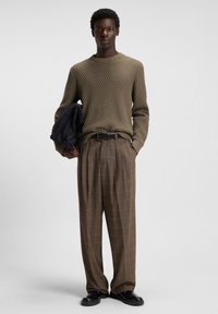 Mann in einem strukturierten olivfarbenen Pullover und braunen karierten Hosen, der eine schwarze Jacke hält und vor einem schlichten hellen Hintergrund steht.
