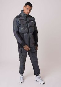 Giacca impermeabile nera e grigia con grandi tasche frontali, colletto alto e cerniera. Abbinata a pantaloni cargo coordinati e sneakers bianche.