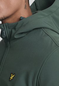 Groene capuchontrui met rits, gemaakt van een zachte stof, met een hoge kraag, contrasterende zwarte stiksels en een klein geel geborduurd logo op de borst.