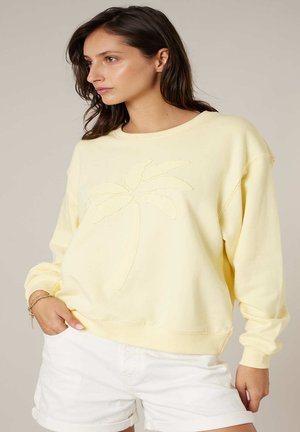 Jeune femme portant un sweat-shirt jaune clair avec un motif de palmier et un short blanc retroussé, debout avec une main dans la poche.