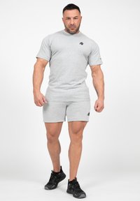 Grijs T-shirt met korte mouwen en bijpassende shorts, beide gemaakt van zacht materiaal. Bevat een klein zwart logo op de linkerkant van de borst en op de shorts. Zwarte sneakers.