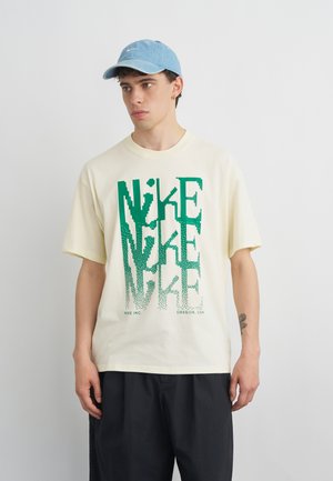 Mladi moški nosi kremno Nike majico z zelenim pikseliziranim logotipom in svetlomodro kapo, stoji pred enobarvnim ozadjem.