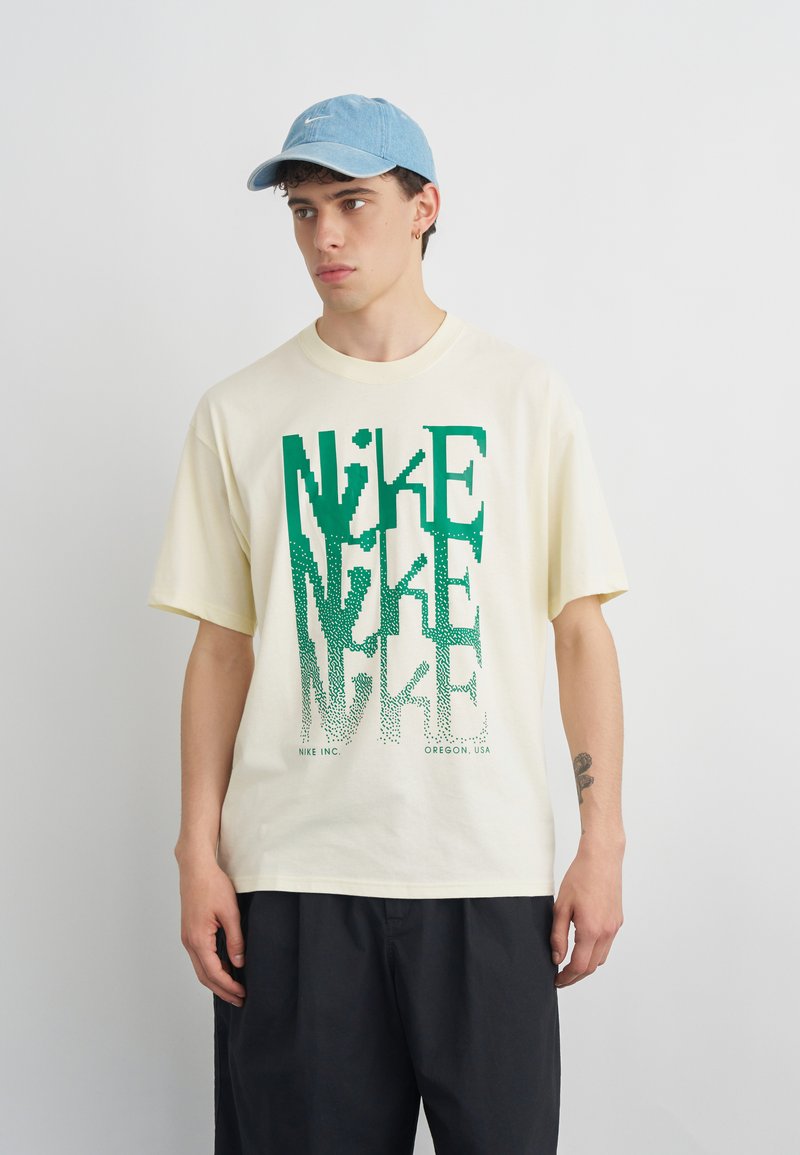 Νέος άνδρας με κρεμ T-shirt της Nike με πράσινο pixelated λογότυπο και ανοιχτό μπλε καπέλο, στέκεται μπροστά σε απλό φόντο.