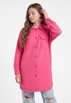 Ung kvinde iført lang, knaplukket, lyserød jakke med brystlommer og stjerneformede, pink øreringe, poserer med hånden nær håret.