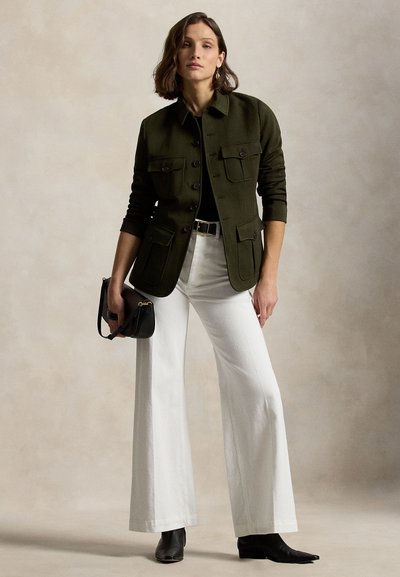 Olijfgroene jas met meerdere zakken aan de voorkant, gecombineerd met een zwart shirt en witte uitlopende broek. Model houdt een kleine zwarte tas vast en draagt zwarte laarzen.