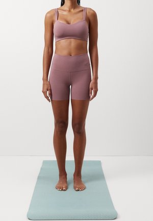 Femme debout pieds nus sur un tapis de yoga bleu, portant un soutien-gorge de sport mauve et un short cycliste taille haute sur un fond blanc uni.