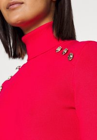 Pull en col roulé ajusté en tissu côtelé rose vif avec trois boutons décoratifs en métal doré sur l'épaule gauche.