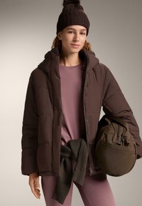 Jachetă matlasată maro cu glugă, cu panouri capitonate. Poartă-o peste un tricou violet deschis cu mâneci lungi, asortată cu o geantă maro.