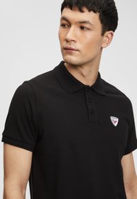 Černé polo tričko s strukturovanou tkaninou, krátkým rukávem a zapínáním na tři knoflíky. Na levé straně hrudi je viditelné malé kulaté logo.