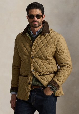 Homme portant des lunettes de soleil foncées, une veste matelassée beige avec col marron, un pull vert et un jean foncé, debout devant un fond uni.