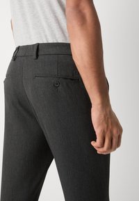 Pantalons gris foncé en tissu texturé. Comprend une poche arrière avec une fermeture à bouton et des passants de ceinture. Placement de la main visible sur la cuisse.