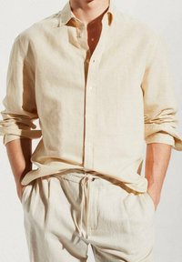 Homme portant une chemise en lin beige clair à manches longues retroussées et un pantalon à cordon assorti, les mains dans les poches.