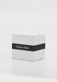 Boîte rectangulaire blanche au fini mat, ornée d'une bande horizontale noire avec l'inscription "Calvin Klein" en texte blanc. Design épuré, sans motifs.