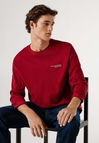 Maglietta a maniche lunghe con scollo rotondo in cotone rosso scuro con testo ricamato bianco. Indossata con jeans blu scuro, seduto su una sedia nera.