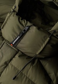 Groene pufferjas met een hoge kraag, voorzien van een ritszak, verstelbare trekkoorden en een merklabel in contrasterende kleuren.