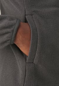 Main partiellement insérée dans la poche latérale d'une veste ou d'un pull en tissu côtelé gris foncé.