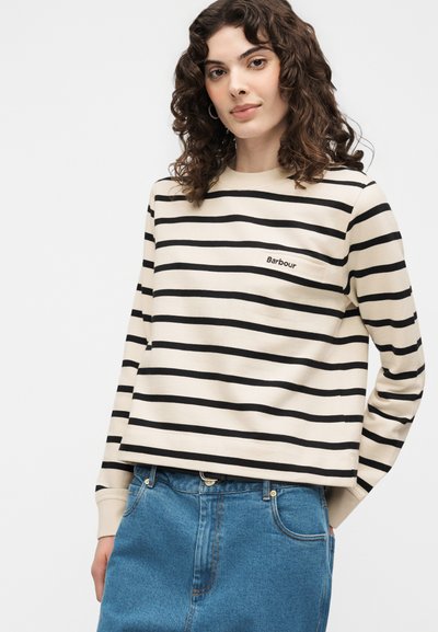 Femme aux cheveux bouclés portant un t-shirt à manches longues rayé crème et noir avec « Barbour » sur la poitrine et une jupe en jean bleue.