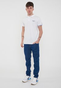 Polo bianco con strisce blu sulle maniche, logo sul petto. Abbinato a jeans blu scuro e sneakers bianche con dettagli blu. Posizione in piedi.