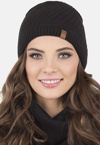 Gorro de punto negro con un patrón texturizado; presenta una pequeña etiqueta de cuero marrón. El modelo que lleva el gorro sonríe, con el cabello largo.