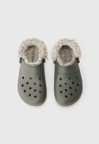 Ein Paar olivgrüne Clogs mit durchlöchertem Obermaterial und beigefarbenem, kuscheligem Futter, entworfen für Wärme und Komfort.