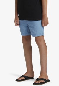 Pantaloni corti di cotone azzurro chiaro con tasche laterali, vita elasticizzata e orlo cucito, abbinati a ciabatte nere su piedi color carne.