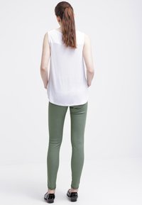 Valkoinen hihaton paita, jossa on upotettu selkäosa, yhdistetty slim-fit oliivinvihreisiin housuihin ja mustiin loafer-kengiin, yksinkertaisen taustan edessä.