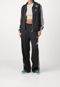 Svart träningsjacka med blomsterbroderi, vita ränder på ärmarna, ihop med svarta byxor, båda i ett mjukt tyg. Turkosa skor fulländar outfiten.