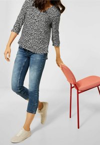 Donna che indossa una blusa nera e bianca con motivo a foglie e jeans blu a vita corta, mentre tiene una sedia corallo-rosa per lo schienale.