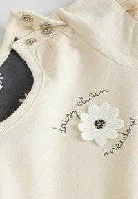 Top de algodón color crema con cuello redondo, botones de presión de metal y una flor blanca bordada con detalles en negro y texto "prado de cadenas de margaritas".