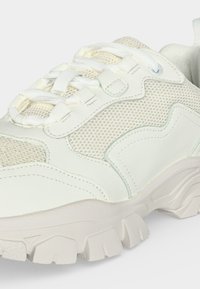 Gros plan sur une chaussure de sport blanche avec tissu en mesh, empiècements en cuir, semelle épaisse et larges lacets noués en boucle.
