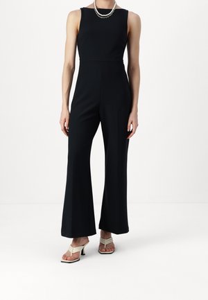 Kvinde iført sort ærmeløs jumpsuit med vide ben, accessoriseret med lagdelte perlekæder og hvide åbne tå-sandaletter med hæl.