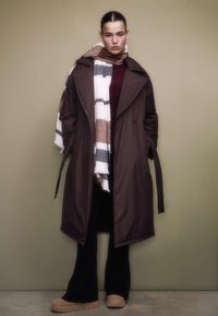 Long brown trench coat with a belted waist, layered over a burgundy top, paired with a striped scarf in white, brown, and grey. Black pants and tan shoes.