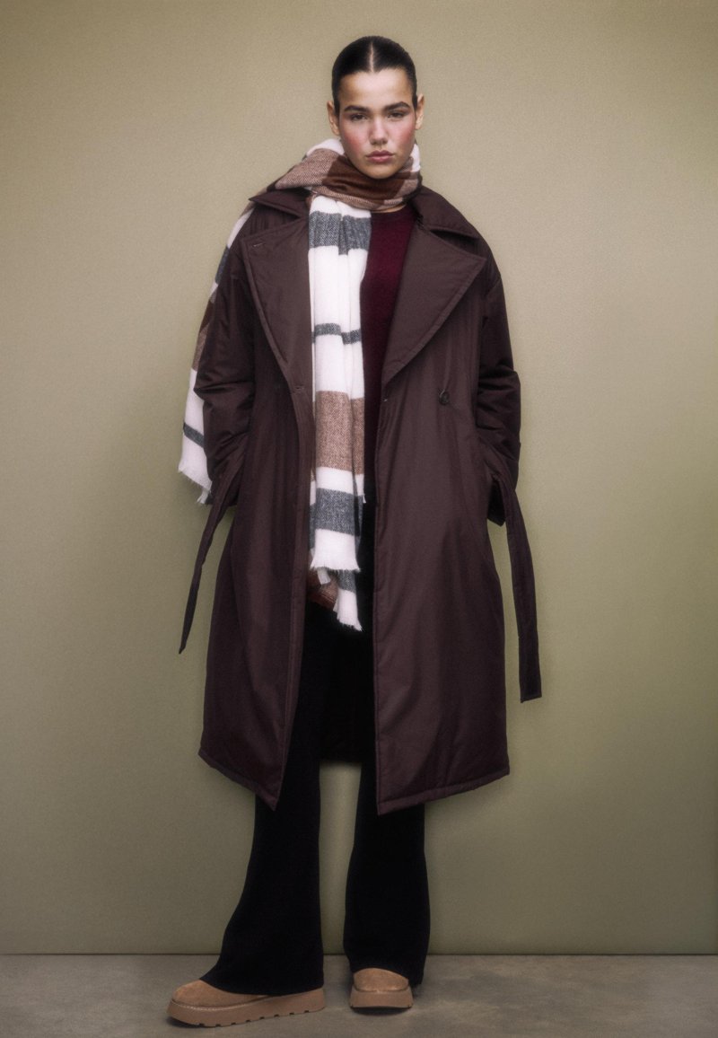 Long brown trench coat with a belted waist, layered over a burgundy top, paired with a striped scarf in white, brown, and grey. Black pants and tan shoes.
