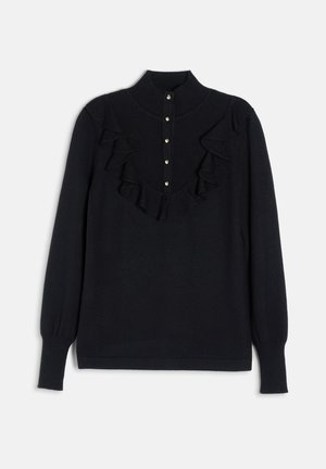 Jonge vrouw met lang donker haar draagt een zwart gerimpeld trui met gouden knopen en een zwarte leren geplooide rok met brede riem.