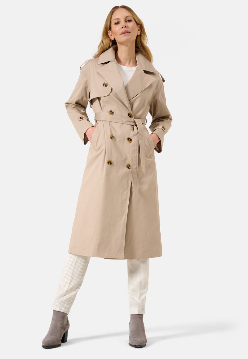 Cappotto beige con design doppiopetto, grandi bottoni e cintura in vita. Modello abbinato a pantaloni a quadri crema e stivali alla caviglia grigi.