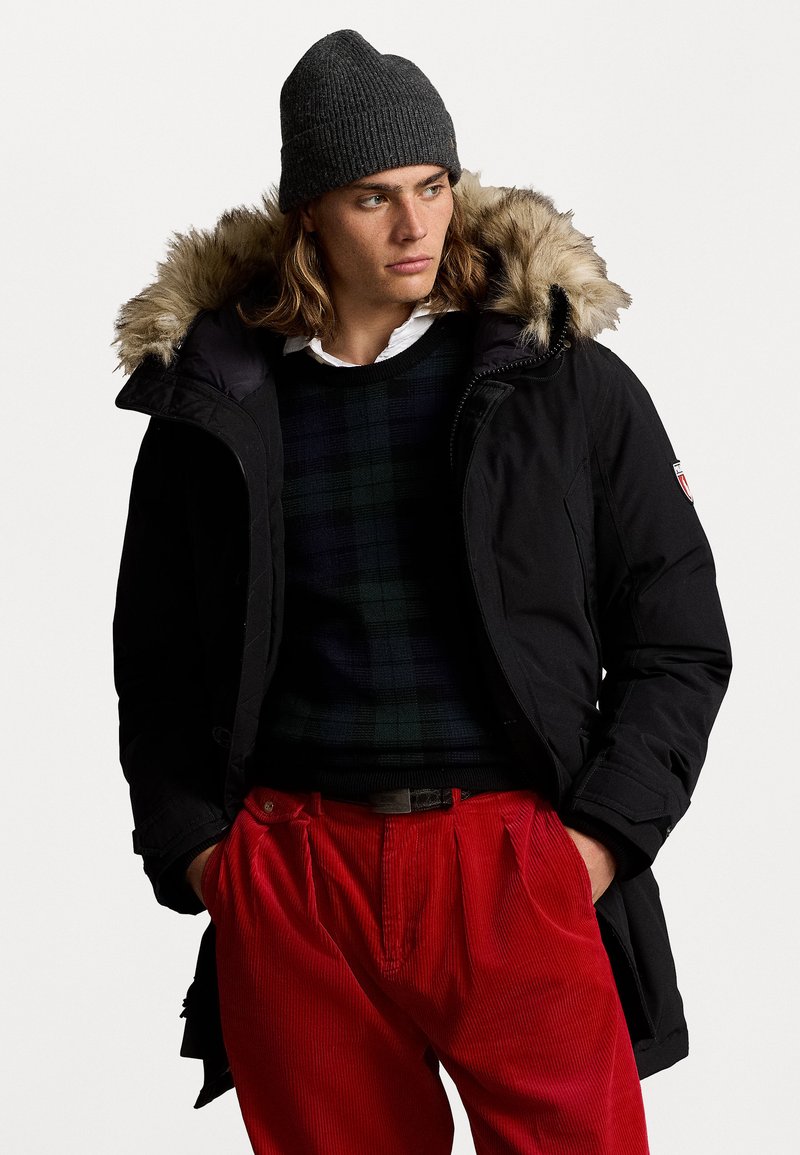 Parka nera con cappuccio foderato in pelliccia, caratterizzata da una chiusura con zip e tasche laterali. Indossata sopra un maglione a scacchi e pantaloni in corduroy rossi.