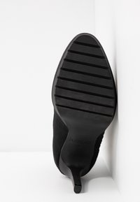 Scarpa nera con tacco alto, dotata di tomaia testurizzata e suola in gomma. Presenta una punta affusolata e scanalature verticali sulla suola per migliorare la presa.
