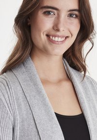 Cardigan grigio a coste con scollatura a V profonda, dotato di maniche lunghe e una texture morbida, indossato sopra una maglia nera. Modello che mostra una vestibilità morbida.