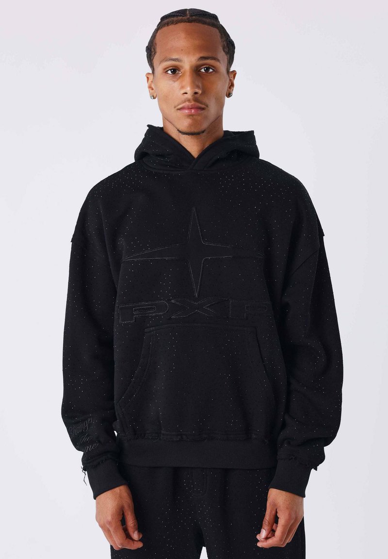 Jeune homme aux cheveux tressés portant un sweat à capuche noir tacheté avec le logo "PXLR" et une étoile, posé devant un fond clair uni.