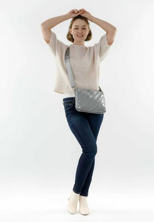 Bolso de hombro acolchado gris con un acabado brillante y correa ajustable, combinado con una blusa de punto ligera y jeans oscuros. Diseño simple y moderno.