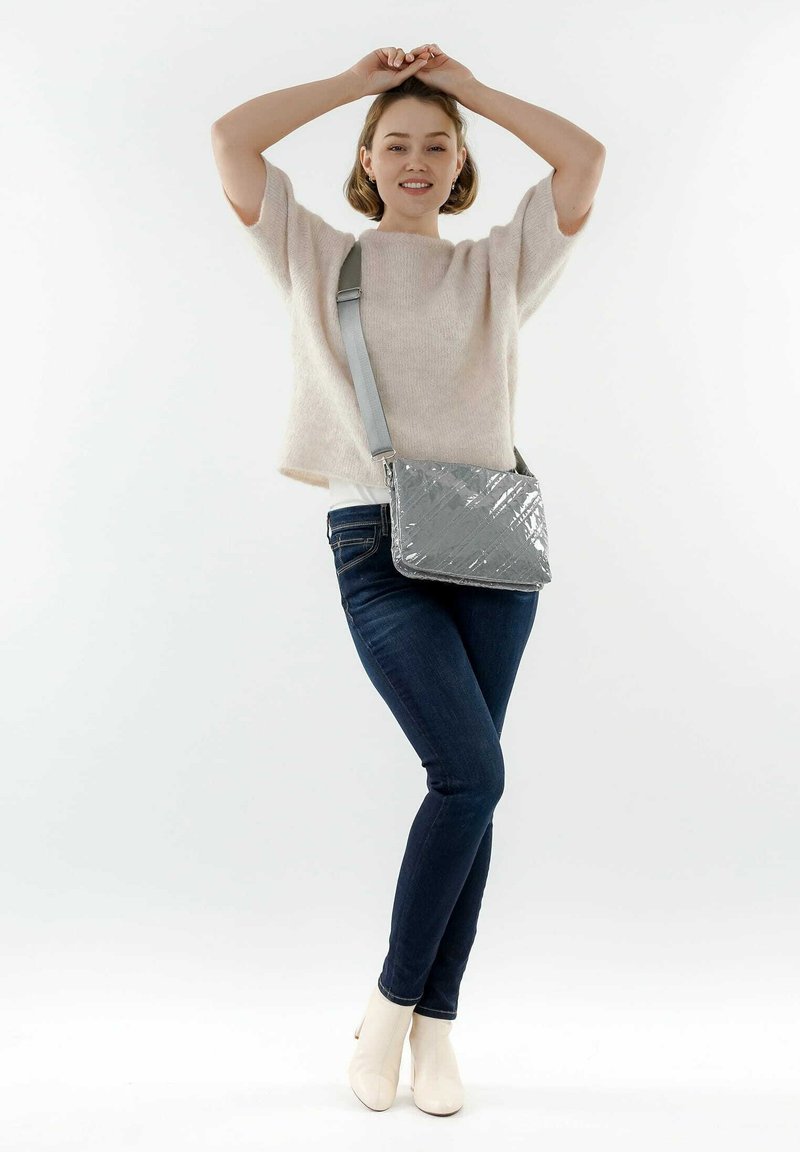 Bolso de hombro acolchado gris con un acabado brillante y correa ajustable, combinado con una blusa de punto ligera y jeans oscuros. Diseño simple y moderno.