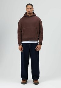 Bruine hoodie met een voorzak, gecombineerd met losvallende donkerblauwe jeans en bruine schoenen. Gladde, casual stoftextuur.