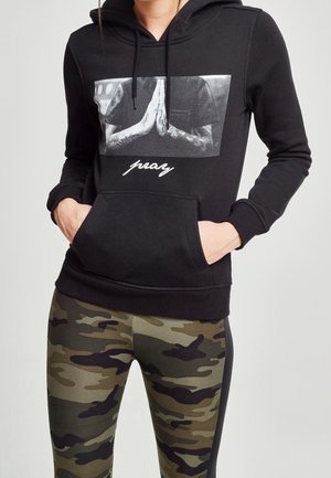 Kvinde iført sort hættetrøje med motiv af bønnehænder og hvid teksten "pray", kombineret med grønne og brune camouflageleggings.