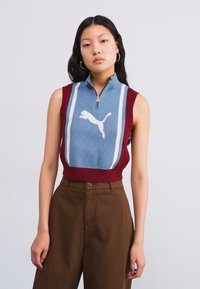 Uten ermer topp har en blå front med hvite striper og Puma-logo, supplert med burgunder sider og glidelås ved halsen.