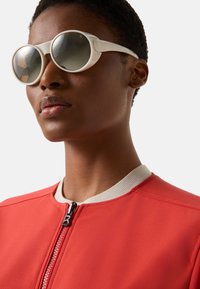 Hvide solbriller med runde linser, en glat ramme og fremtrædende hardware. Båret med en rød bomberjakke med en kontrasterende krave.