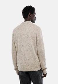 Pull en tricot de couleur crème avec une surface texturée, présentant des éclats de brun, de noir et d'orange ; ourlet et poignets à côtes.