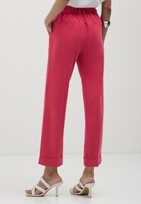 Persona che indossa pantaloni larghi di un brillante colore rosa con orli a coste e sandali bianchi con tacco alto, in piedi con le mani in tasca.