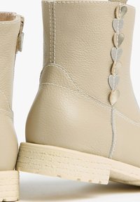 Beige leather ankle boots with textured finish, featuring silver heart-shaped accents along the side and a rubber sole.